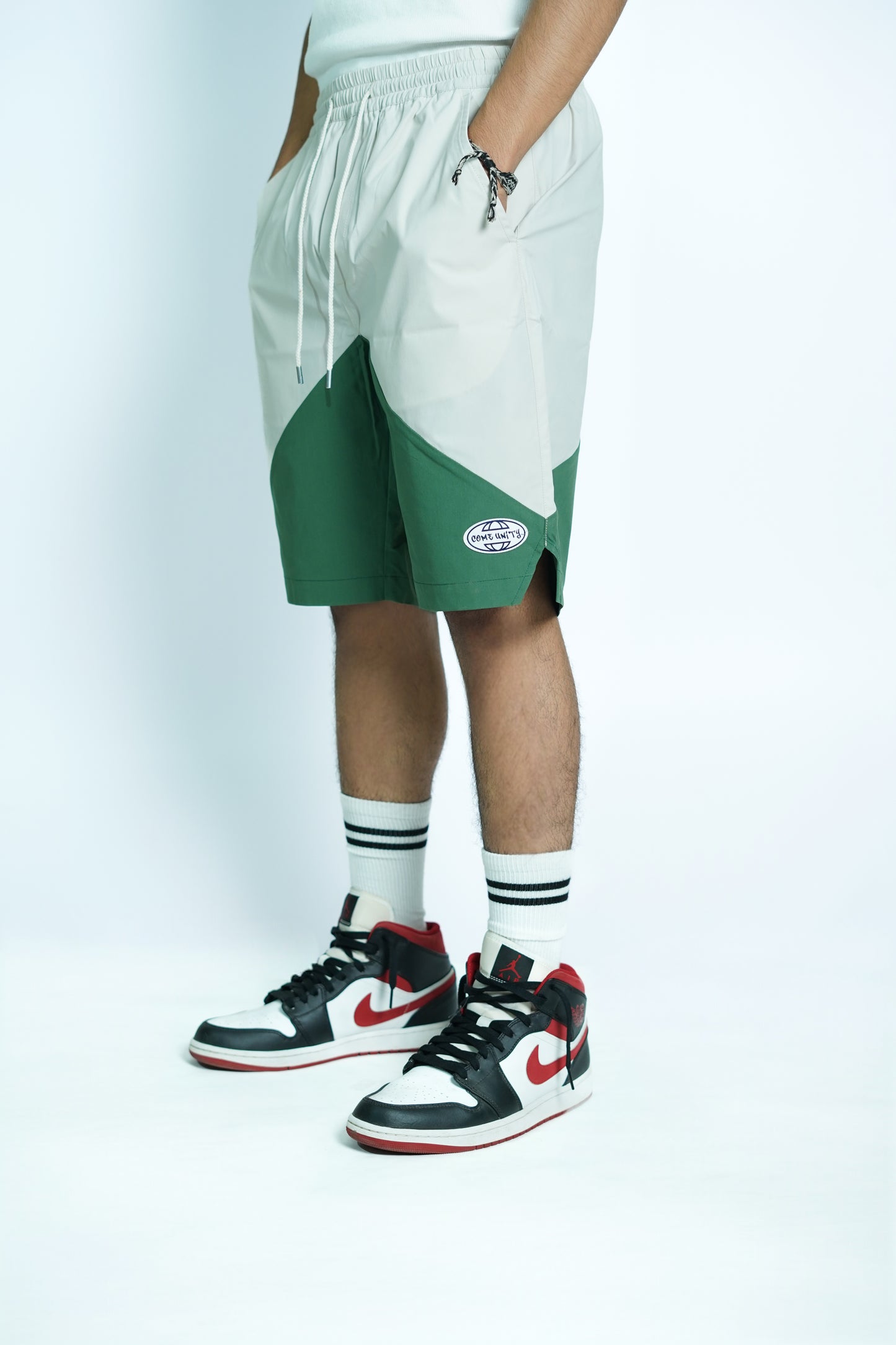 Person wearing white and green shorts with a COEMUNITY logo, black and red sneakers, and striped socks on a light gray background.