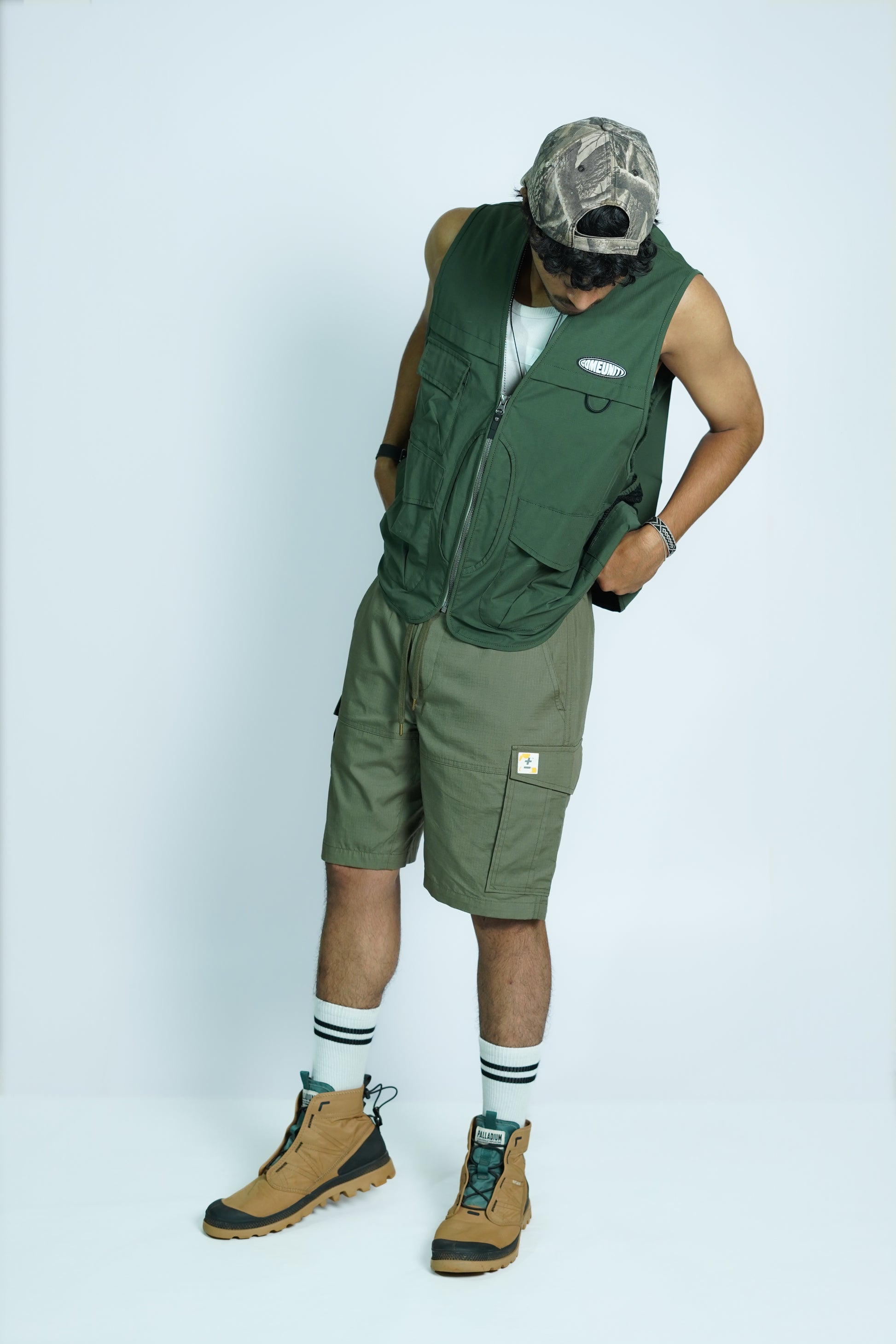 Person wearing COMEUNITY's green sleeveless vest and shorts with a camouflage cap on a white background
