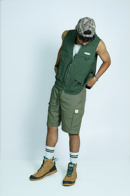 Person wearing COMEUNITY's green sleeveless vest and shorts with a camouflage cap on a white background